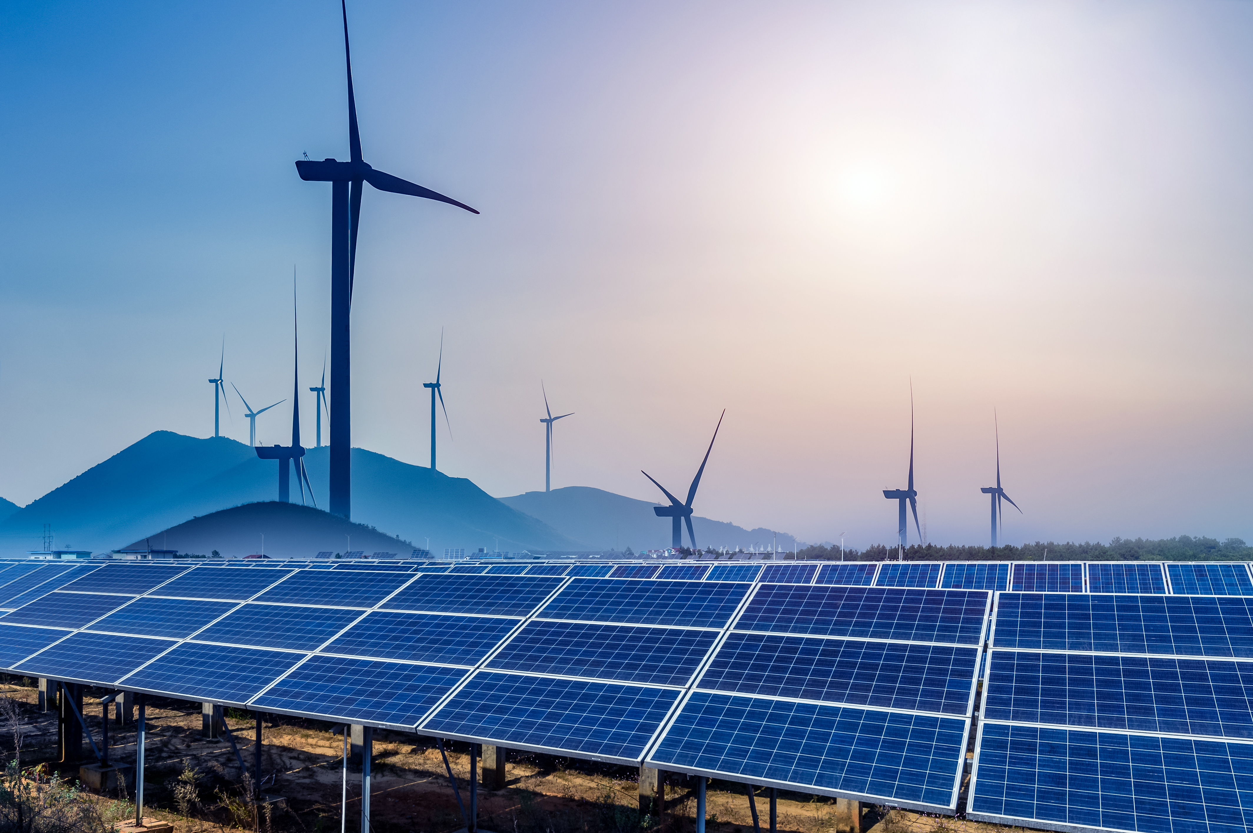 Vor Windrädern auf einer Hügelkette reihen sich Solarmodule auf einer Wiese.