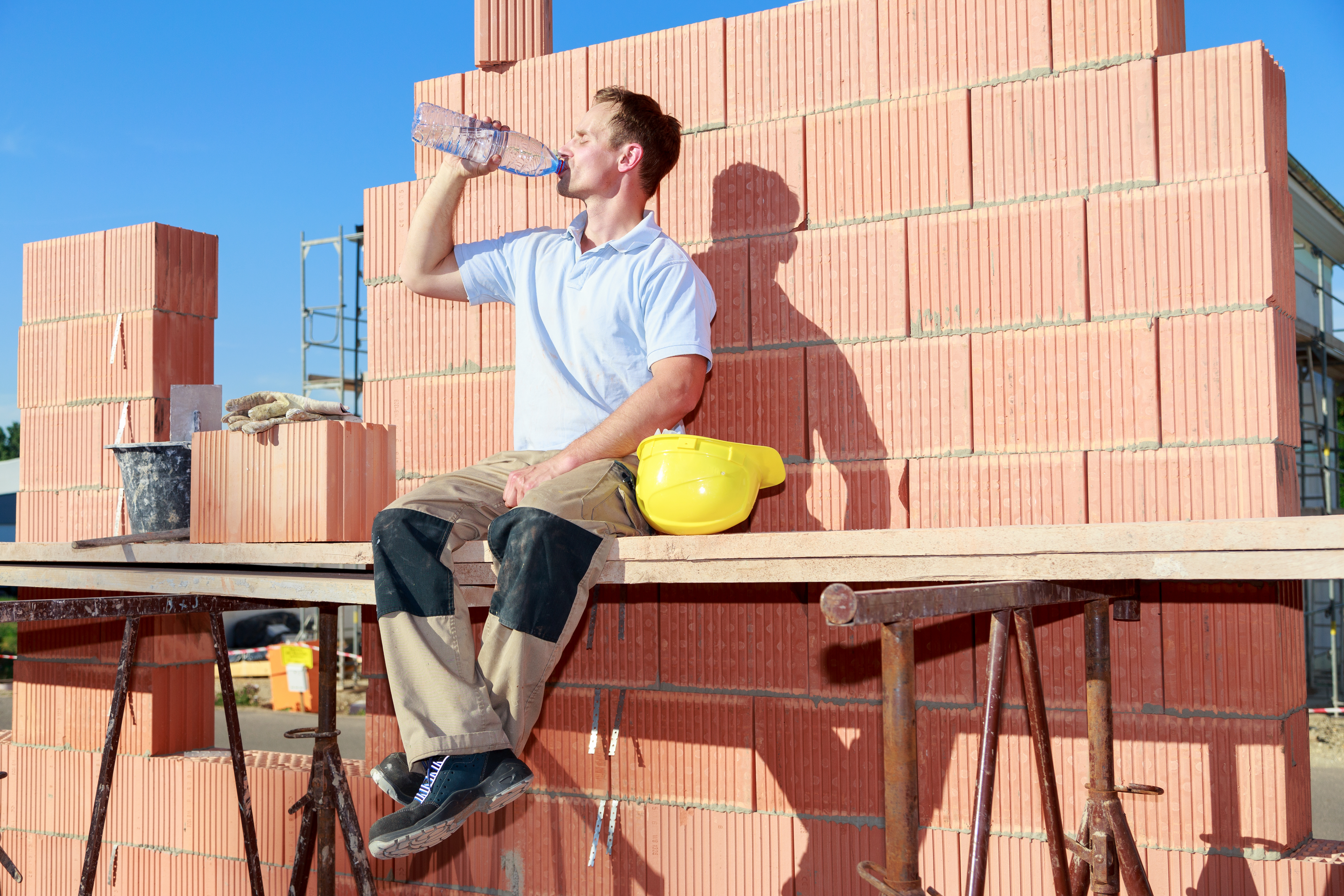 Bauarbeiter sitzt bei starkem Sonnenschein vor einer Mauer und trinkt Wasser.