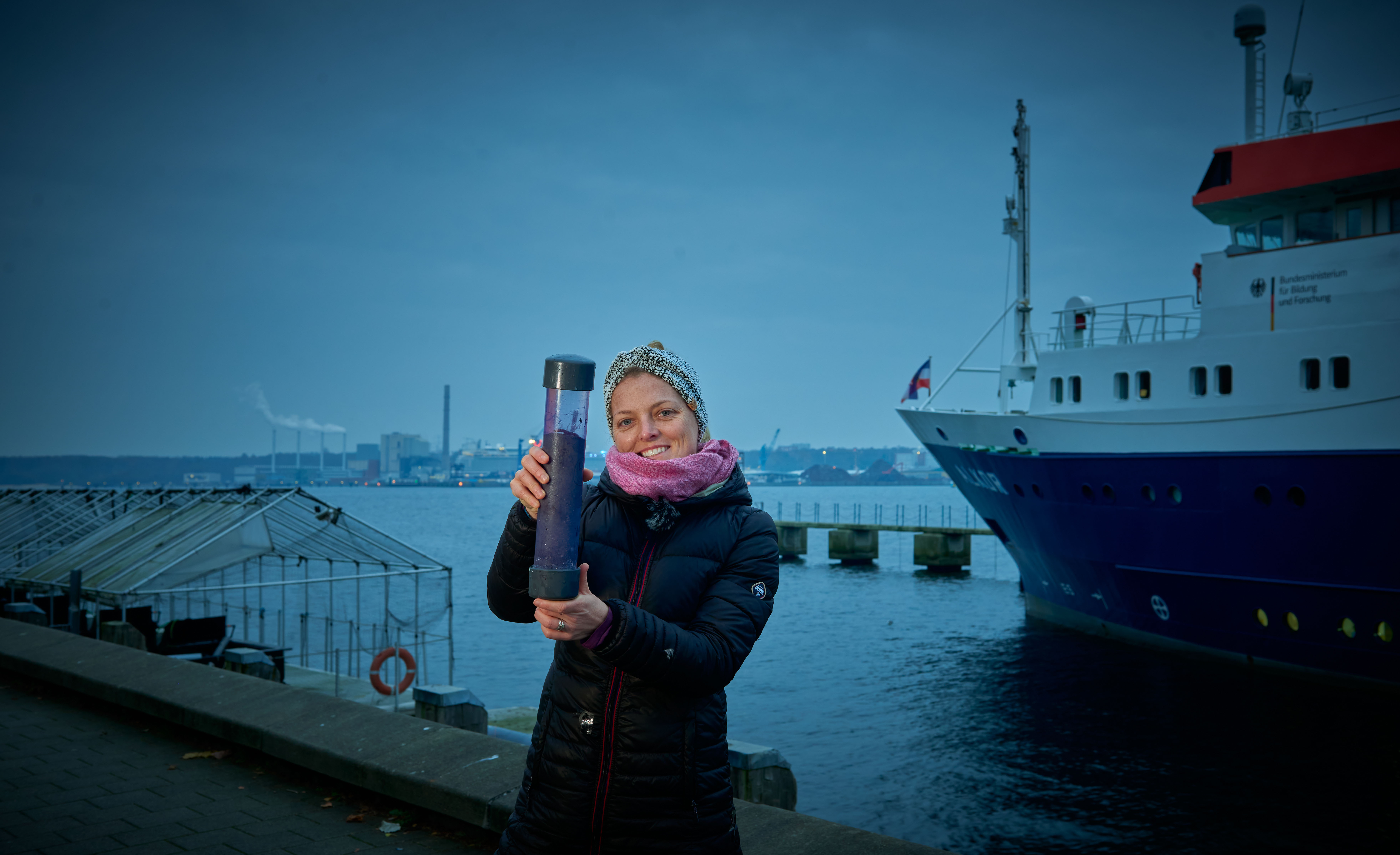Angela Stevenson steht im Morgengrauen in einem Hafen und hät einen Plastikzylinder mit Sediment in die Kamera. Im Hintergrund sient man ein Schiff.