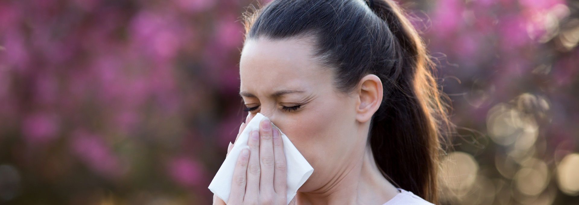 A person blowing their nose, with flowers and trees in the background