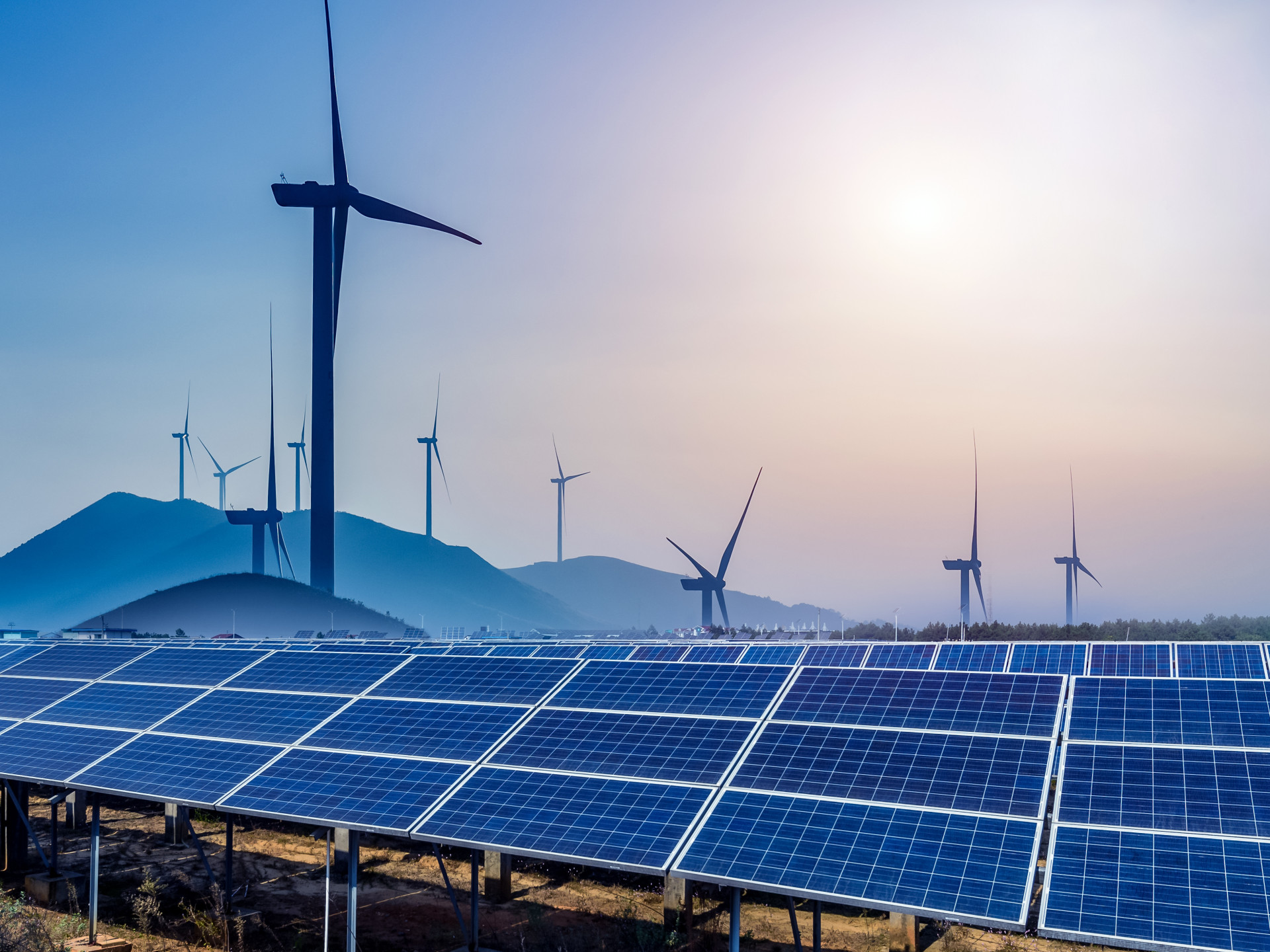 Vor Windrädern auf einer Hügelkette reihen sich Solarmodule auf einer Wiese.