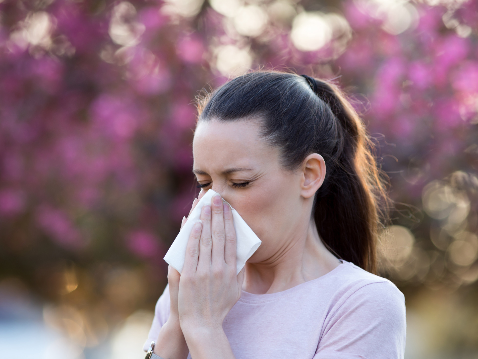 A person blowing their nose, with flowers and trees in the background