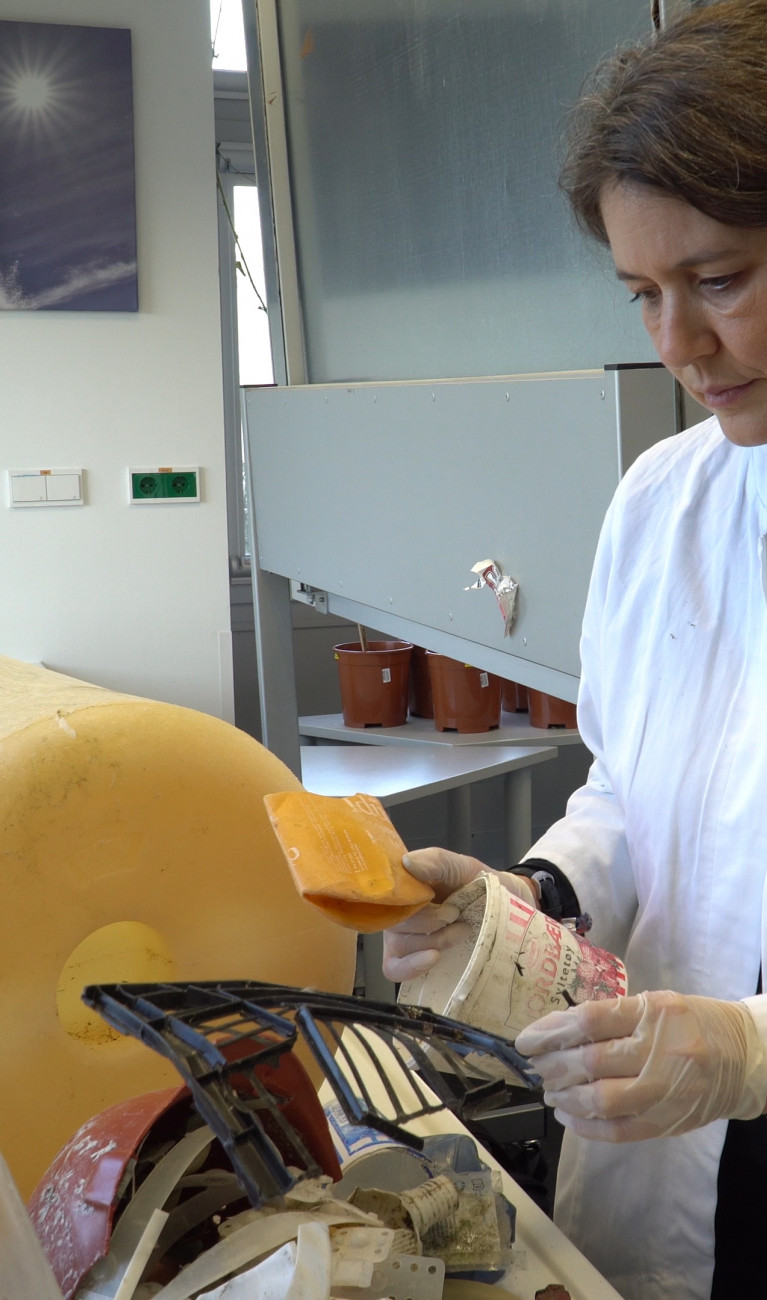 Wissenschaftlerin im weißen Kittel mit einem großen gelben Stück Plastikmüll im Labor