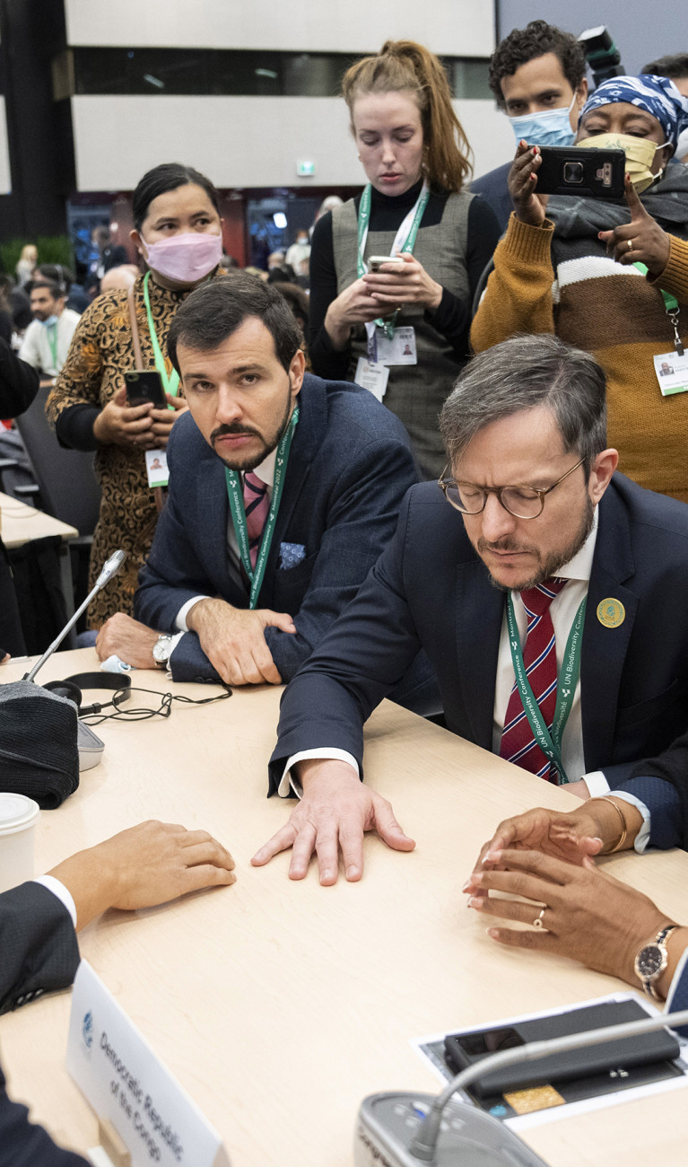 COP15 participants from different countries sitting at a table negotiating, and being photographed from people standing in the background.