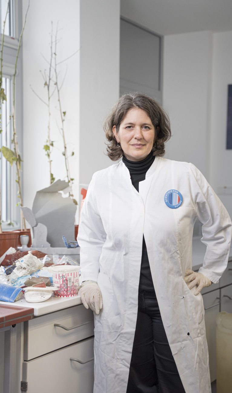 Scientist Melanie Bergmann in a white lab coat in front of a collection of plastic waste