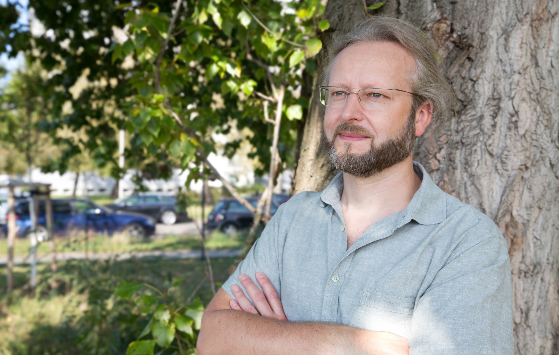 Portrait Björn Rau steht vor Baum, trägt ein grünes Hemd. Arme verschränkt.