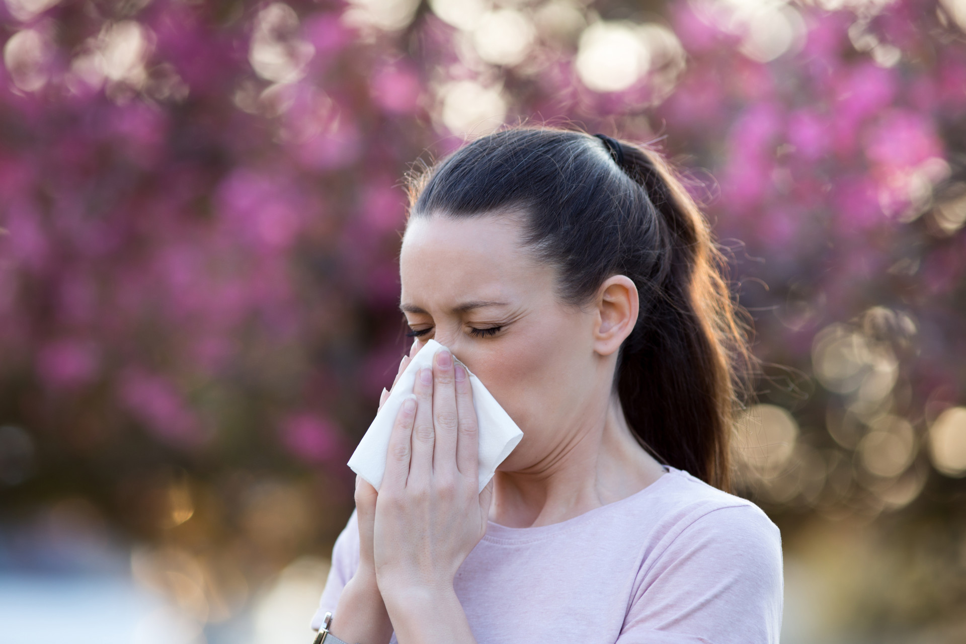 A person blowing their nose, with flowers and trees in the background