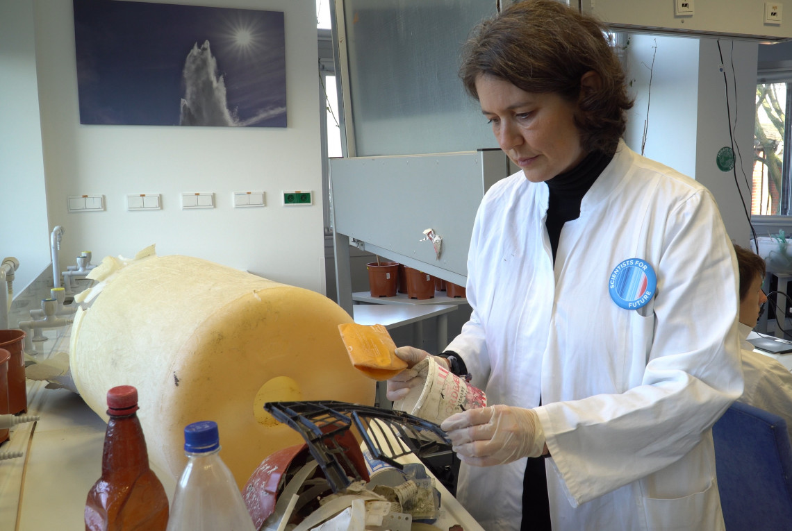 Wissenschaftlerin im weißen Kittel mit einem großen gelben Stück Plastikmüll im Labor