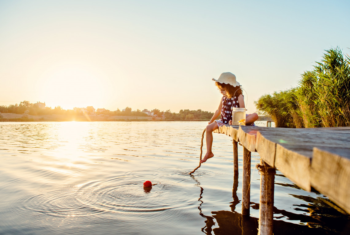 Es ist Sonnenuntergang. Ein kleines Mädchen sitzt auf einem Holzsteg und hält eine Angel in den See.