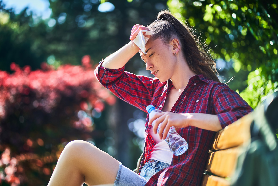 A young woman sits on a park bench. She is sweating and drinking from a water bottle.