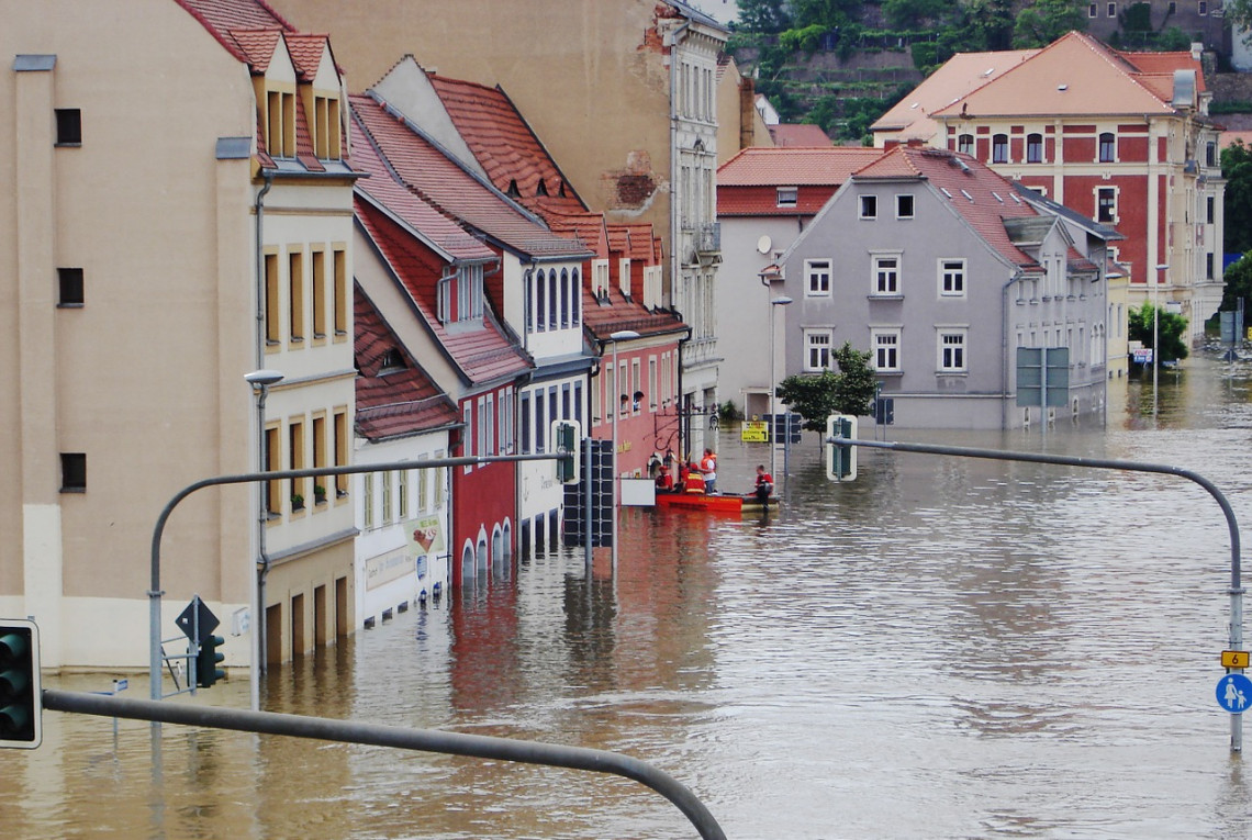 Eine Straße steht komplett unter Wasser, Menschen in einem Schlauchboot retten Personen aus einem Haus